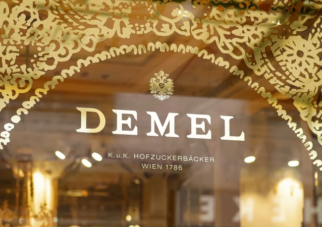 Ornate gold lettering spells 'Demel' on a shop window, with intricate gold detailing above and below the text in a warm, inviting setting.