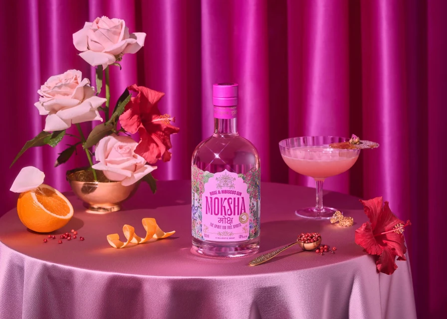 Artful beverage staging featuring a Moksha gin bottle, cocktail glass, hibiscus flowers, roses, and orange on a pink tablecloth.