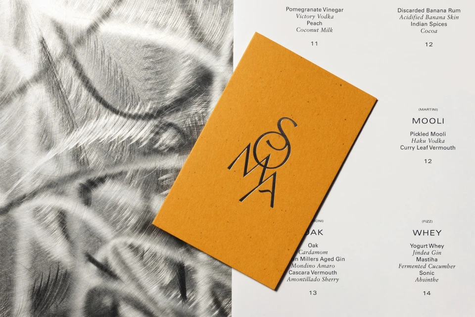 A stylish cocktail menu featuring NOMA's logo is displayed against a brushed steel surface and a white background with drink descriptions.