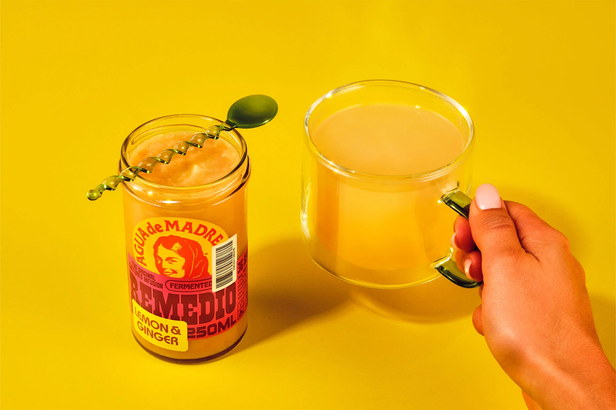 Glass mug and jar with AGUA de MADRE fermented lemon ginger drink with green spoon on yellow background in food and beverage still life.