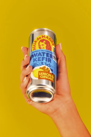 A hand holds a silver can of Agua de Madre Water Kefir Lemon & Ginger drink against a bright yellow background in a studio shot.