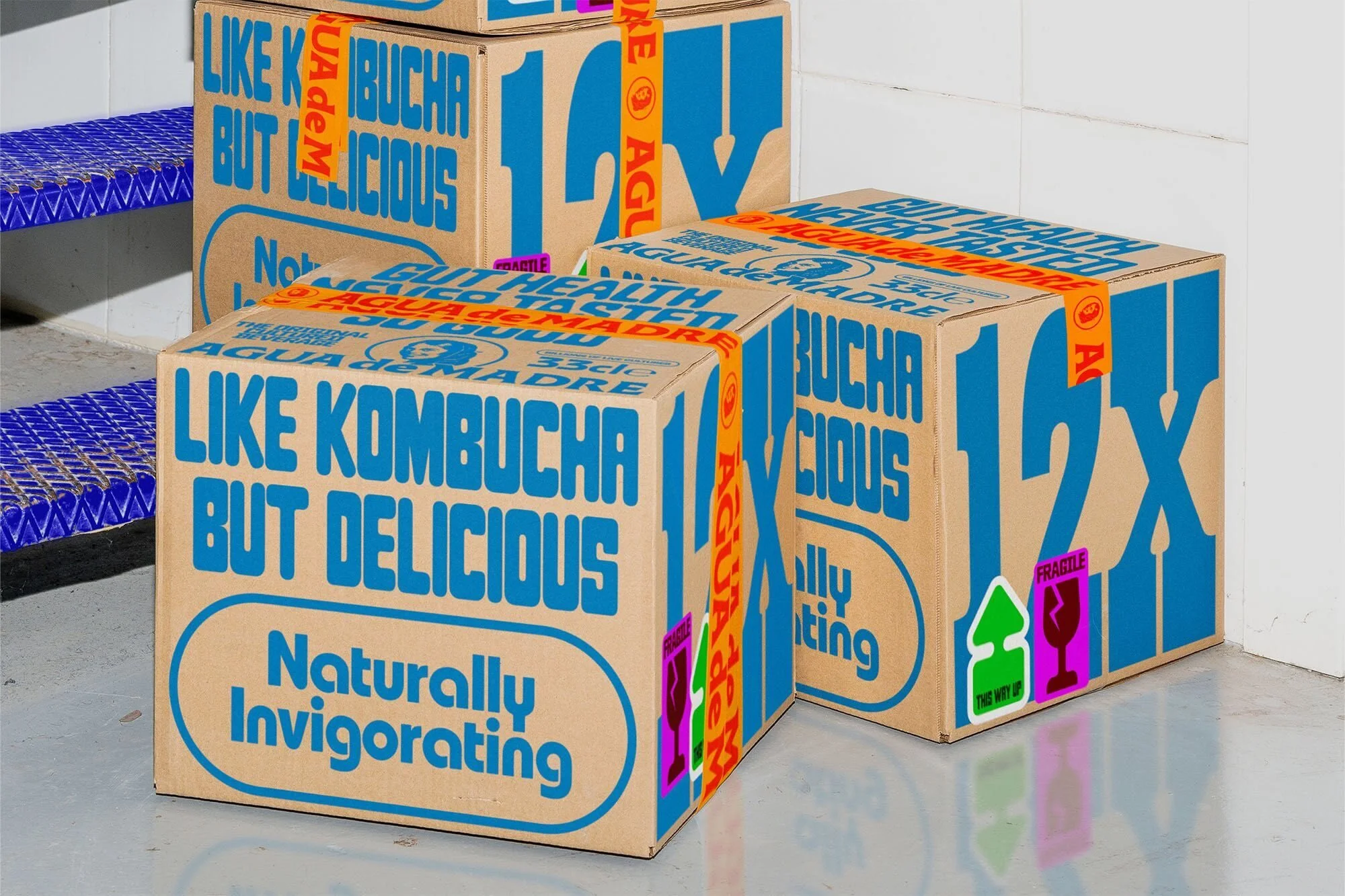 Several cardboard boxes with blue text and tape, stacked in an indoor setting with a tiled floor and blue metal steps visible in the background.