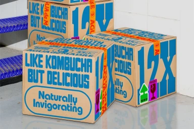 Branded Agua de Madre cardboard shipping boxes with blue typography and orange tape stacked in a room.