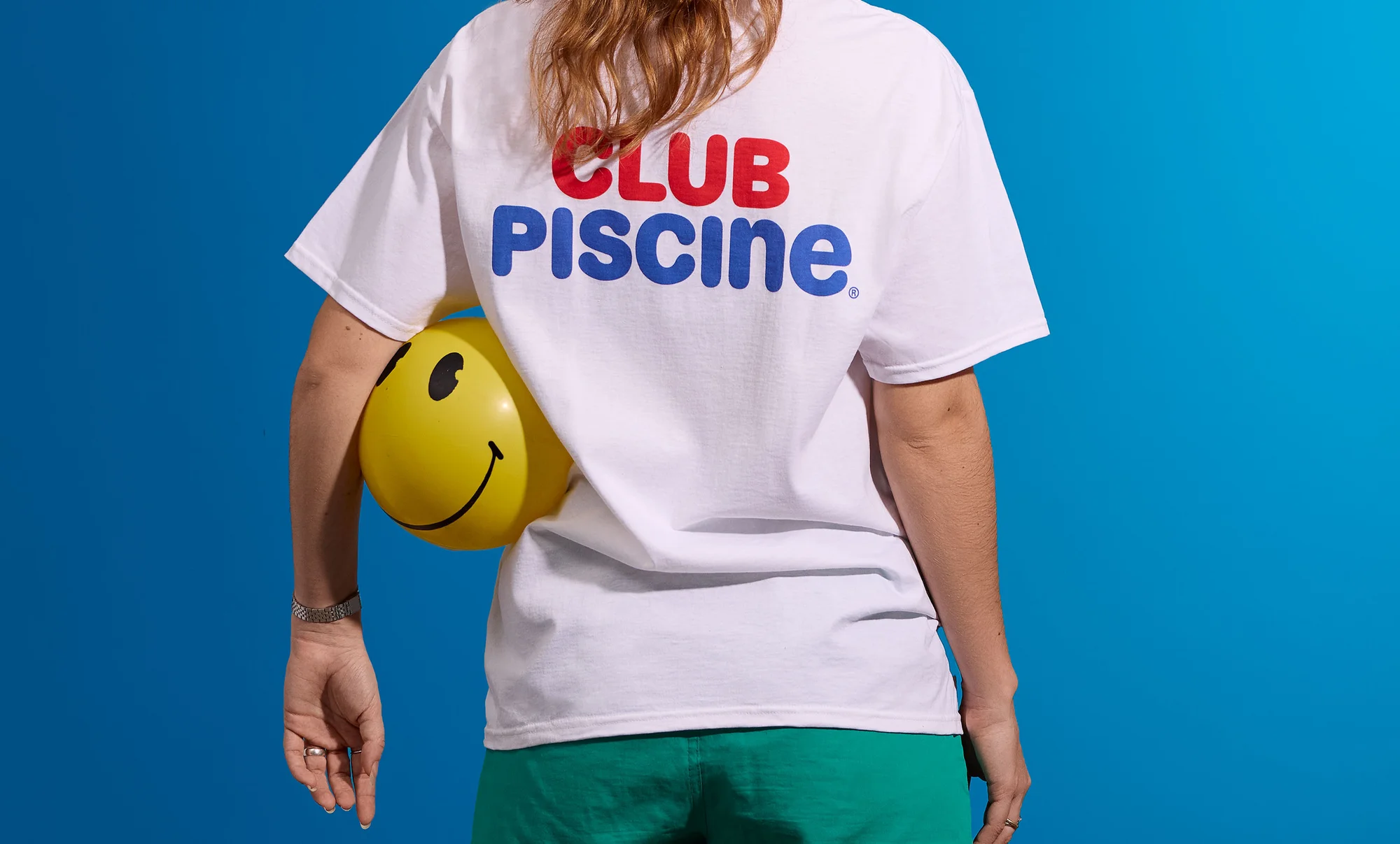 Back view of an adult wearing a white t-shirt with 'Club Piscine' and holding a yellow smiley-face balloon against a blue backdrop.