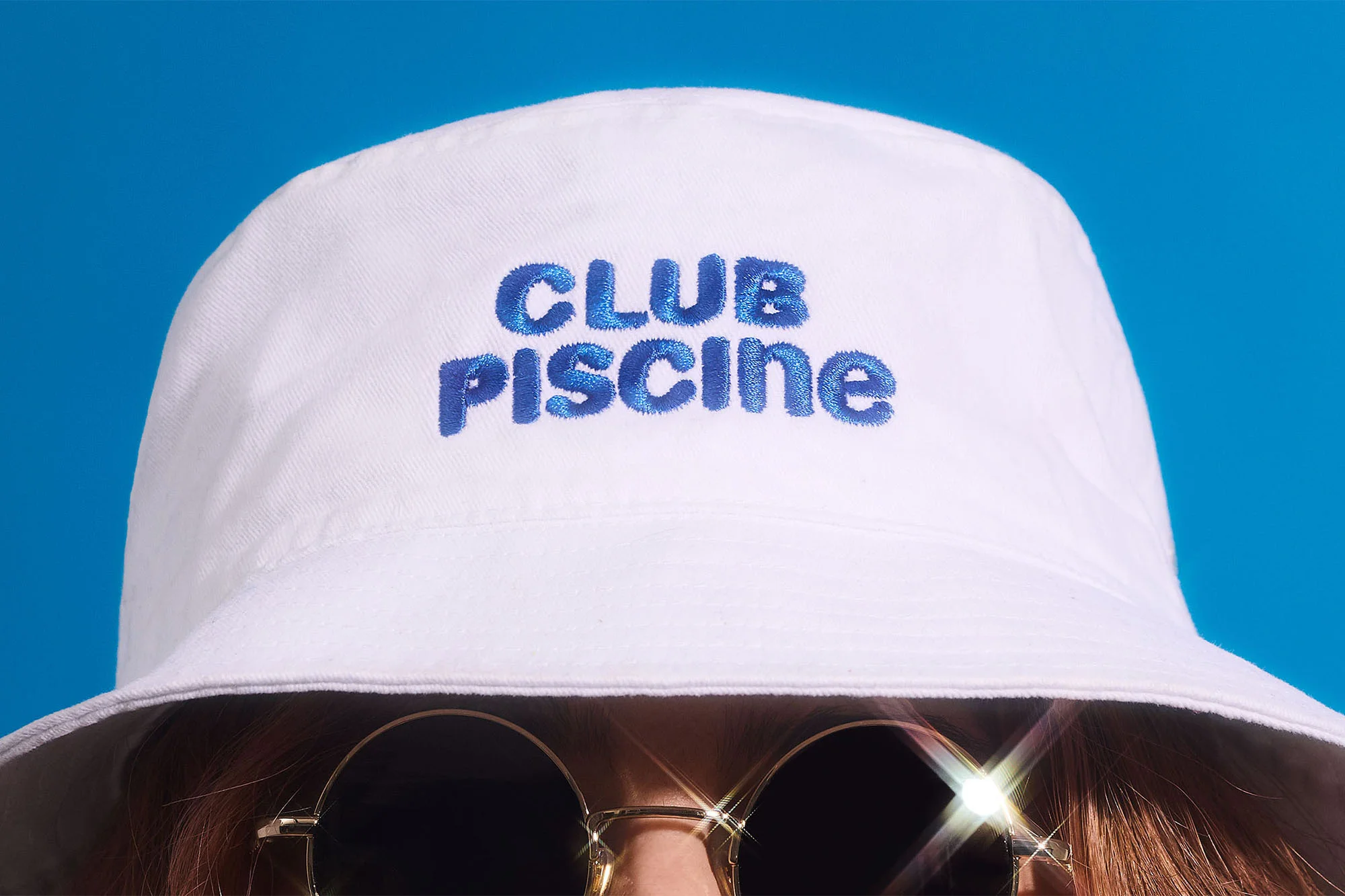Person wearing a white bucket hat with 'CLUB PISCINE' in blue embroidery and round sunglasses, set against a blue background.