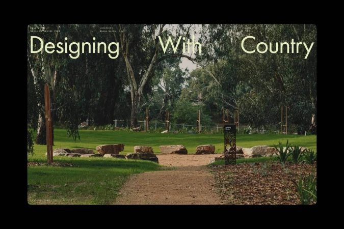 Exterior daytime view of a park featuring a lawn, trees, a pathway, and boulders. A sign provides information about the location.