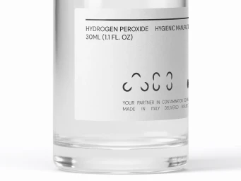 Close-up product shot of a clear glass bottle with a minimalist white label featuring typography and graphic icons against a bright white background.