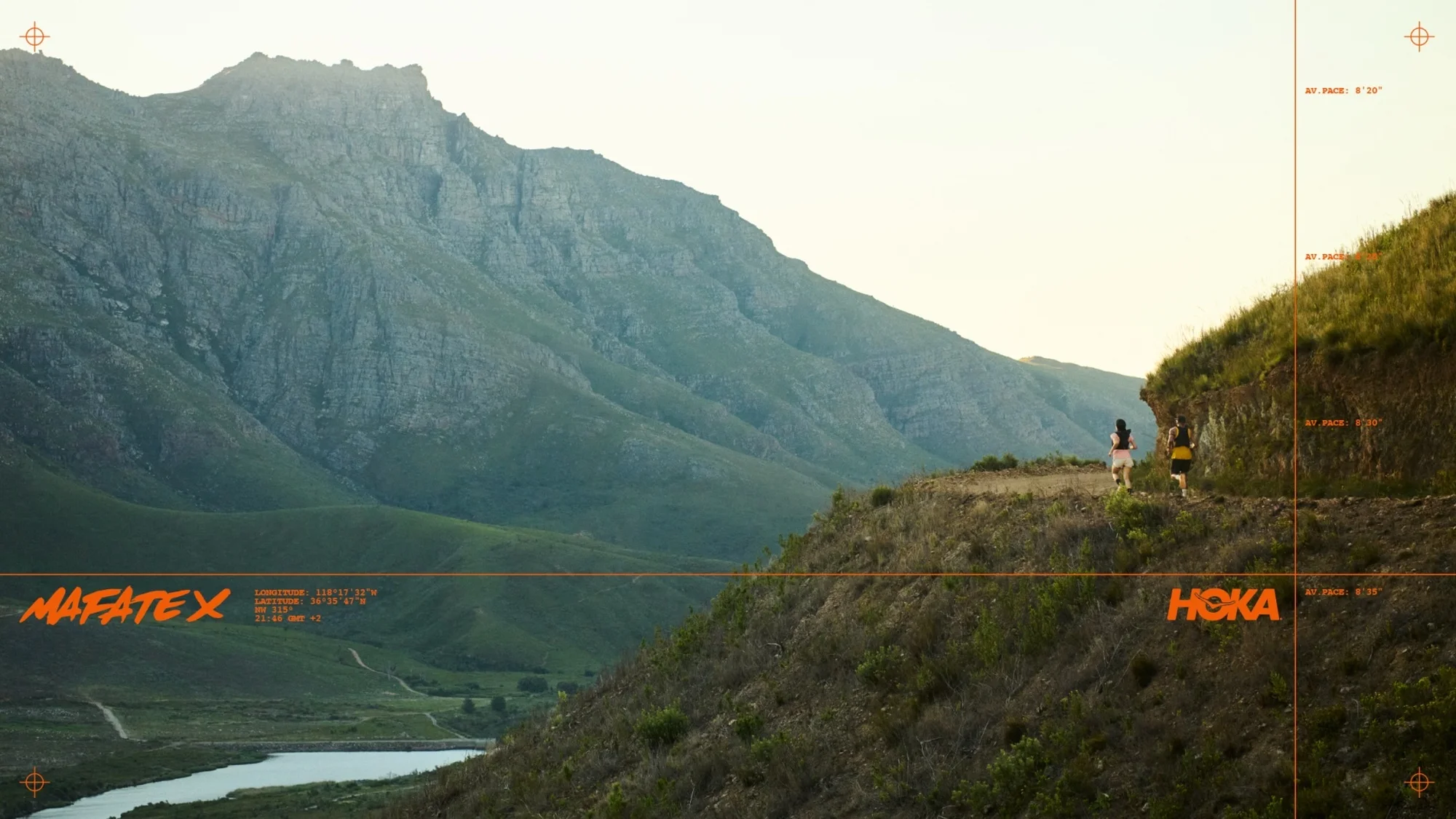 Two people trail running on a dirt path along a green hillside, with a mountain range in the background under a clear sky, athletic wear.