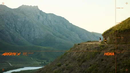 Two runners on a high dirt path through a mountainous landscape with digital orange graphic overlays and HUD elements.