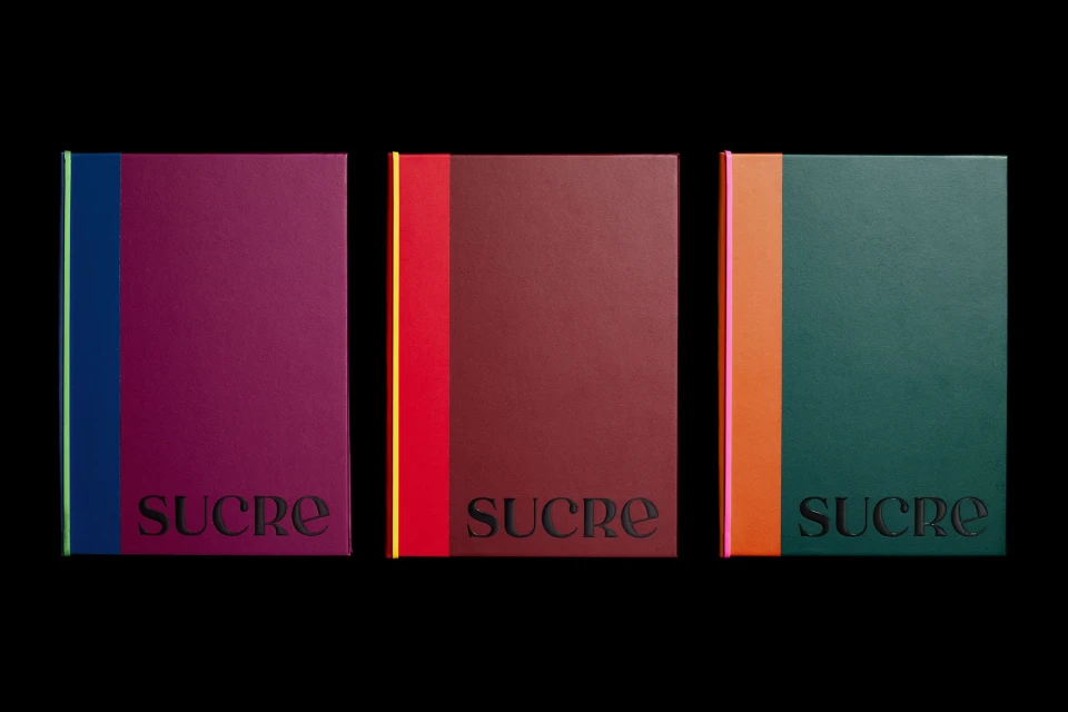 Three hardcover books with a vertical stripe on the left side, displayed against a black background. Each book has the word 'SUCRE'.