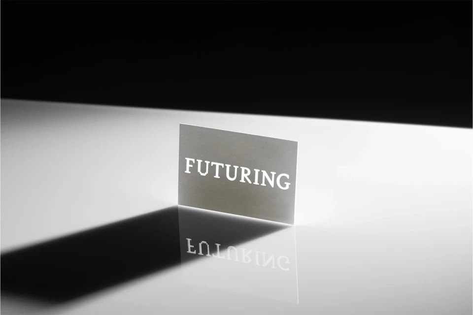 Abstract studio shot of a backlit card with the text 'FUTURING' cut out, casting a sharp shadow and reflection on a glossy surface.