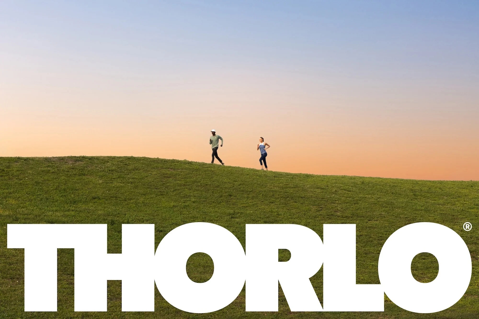 Two athletic adults jog on a grassy hilltop at sunset, with the large white 'THORLO' logo in the foreground, creating a vibrant, active scene.