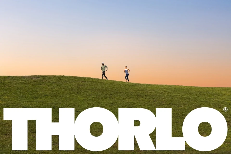 Two athletic adults jog on a grassy hilltop at sunset, with the large white 'THORLO' logo in the foreground, creating a vibrant, active scene.