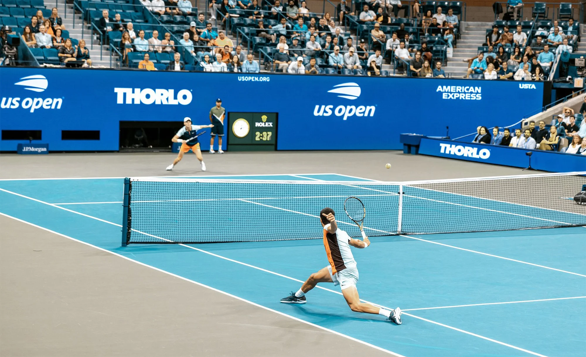 A tennis match on a blue court features two male players mid-game, with spectators in the background and visible sponsor signage.