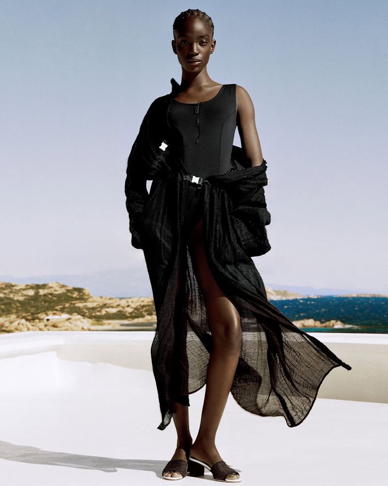 Fashion portrait of a Black woman in a black swimsuit and cover-up, posing outdoors with a coastal background on a sunny day.