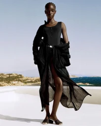 Model standing outdoors against a coastal background wearing a black swimsuit and sheer black wrap