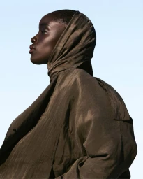 Profile portrait of a Black female model wearing an olive green hooded coat against a clear pale blue sky.