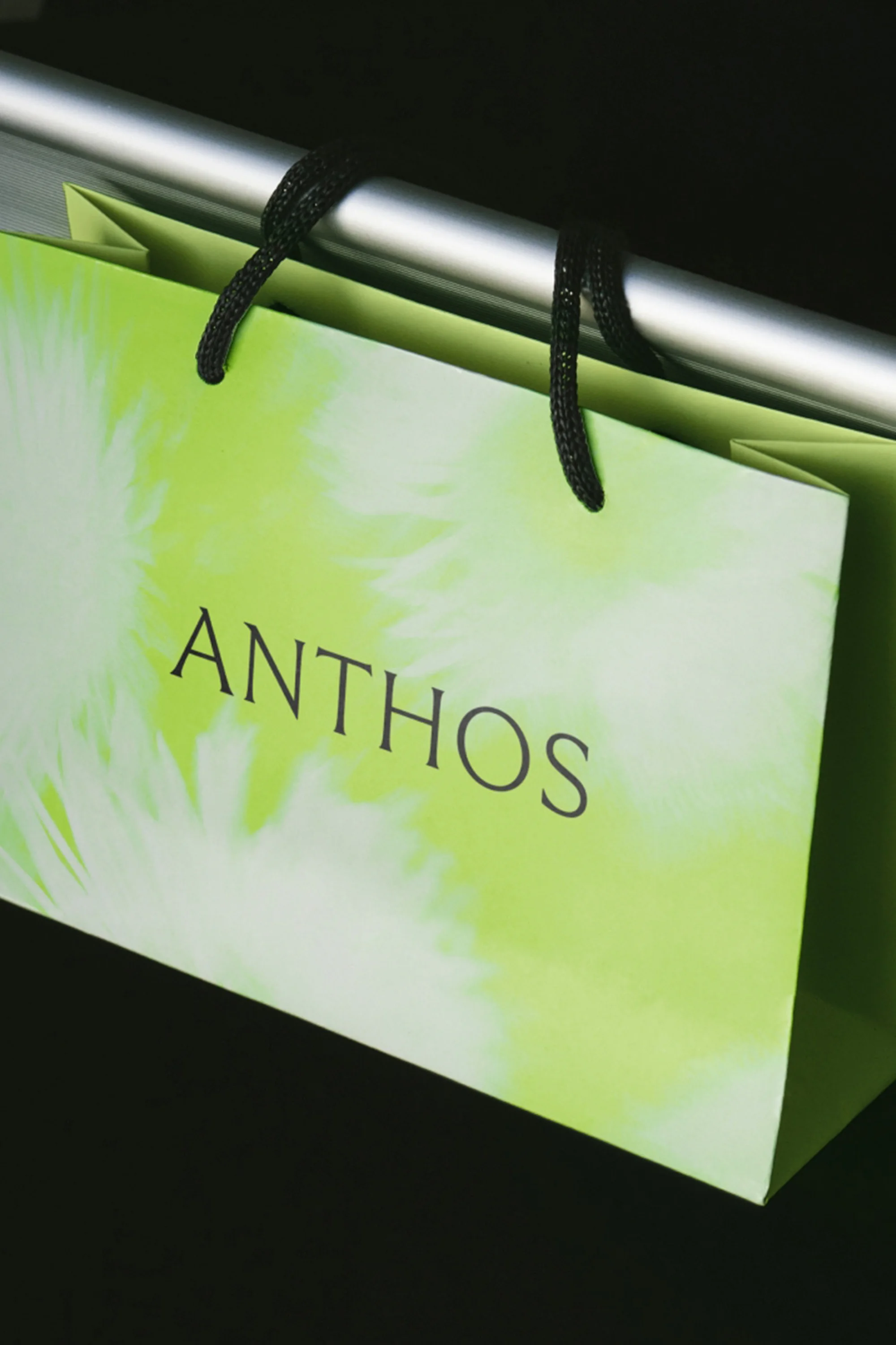 Close-up studio shot of a green Anthos shopping bag with black rope handles hanging on a silver metal bar against a dark background.