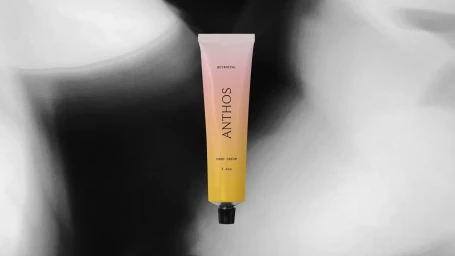 Gradient yellow and pink hand cream tube with black typography centered vertically against a blurred black and white background.