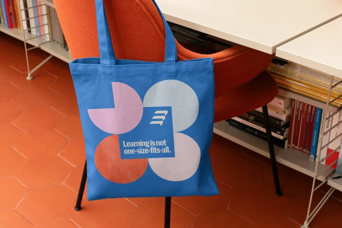 Blue tote bag with colorful graphic design on orange tiled floor. An orange chair and white shelves are in the blurred background.