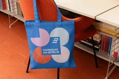 A blue canvas tote bag featuring geometric shapes and the text Learning is not one-size-fits-all hanging on an orange chair.