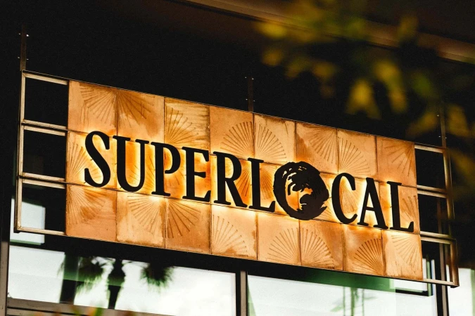 Illuminated 'SUPERLOCAL' sign hanging outside a building, featuring modern font and design, with foliage in the background. Sign