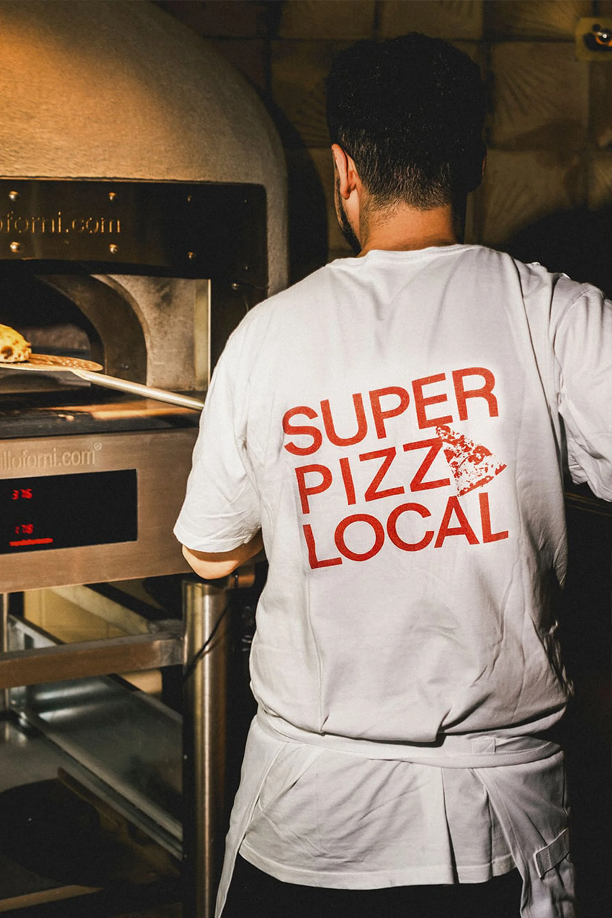 Adult male employee putting a pizza inside a pizza oven inside a restaurant, wearing a white t-shirt with the text SUPER PIZZ LOCAL.