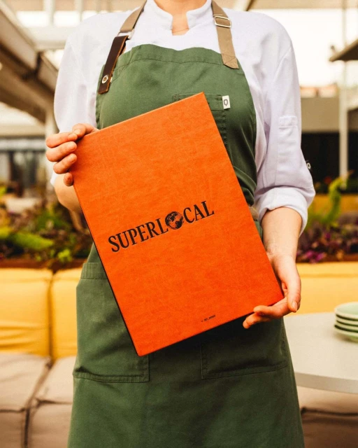 Chef presents an orange menu with the text 'SUPERLOCAL' while wearing a green apron, offering a glimpse into the culinary world.