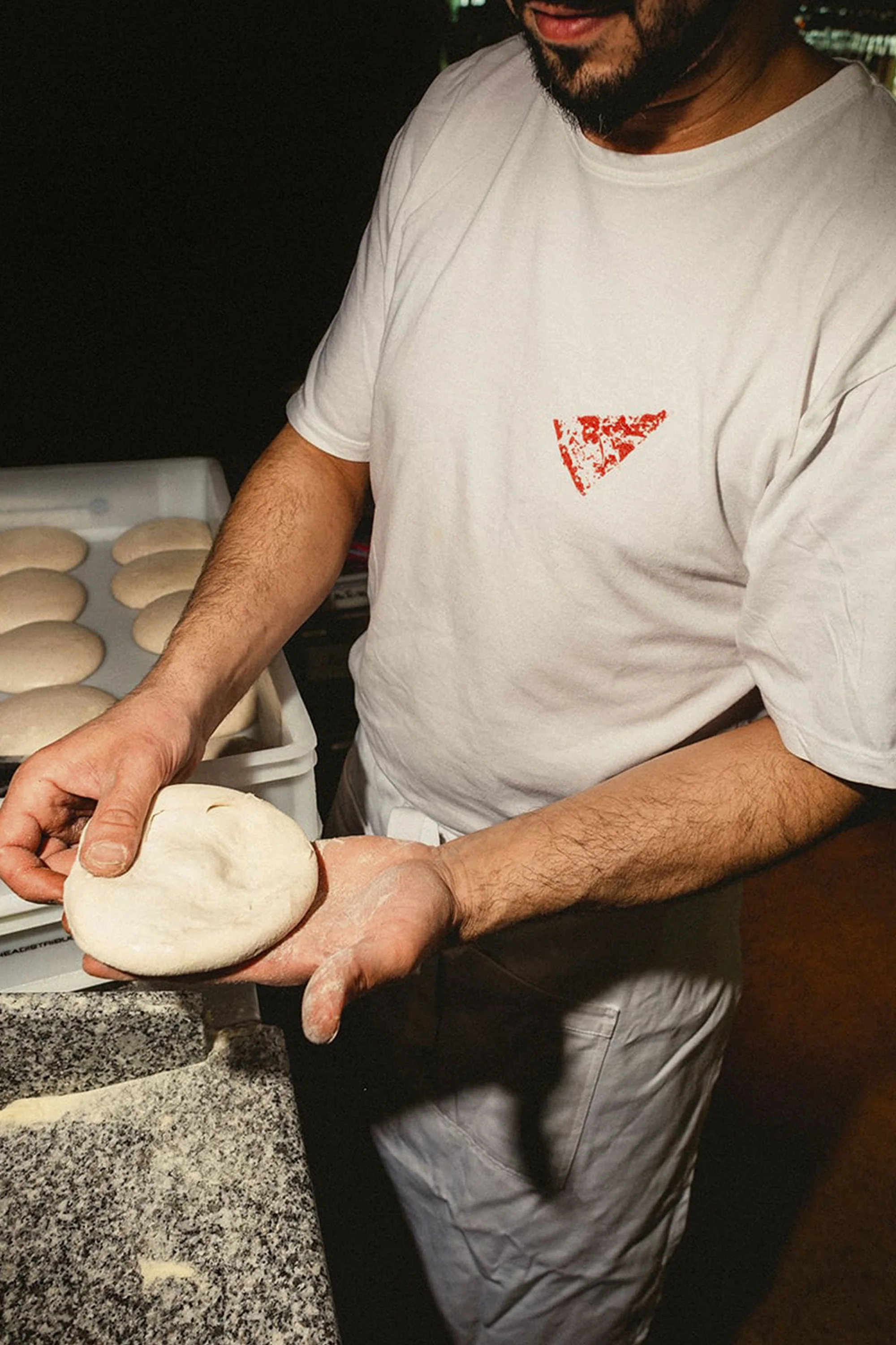 Adult male chef prepares round pizza dough in his hands in an indoor restaurant kitchen with trays of dough in the background.