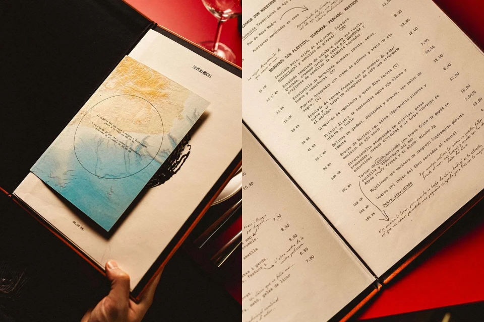 An open restaurant menu displays Spanish text, set against a red table surface with a wine glass, featuring a neutral-toned card.