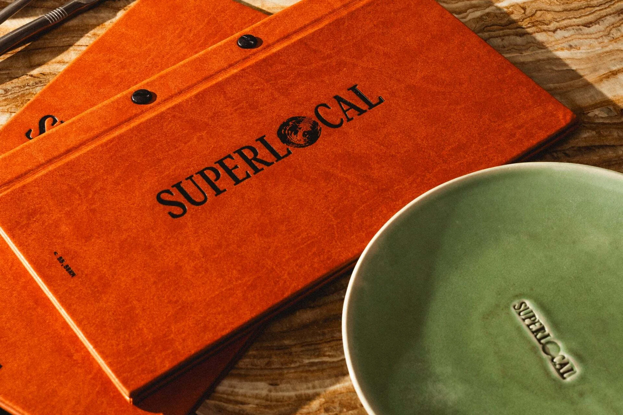 Close-up of orange menu books with 'SUPERLOCAL' text, resting on a marble surface next to a green ceramic plate with the same text.