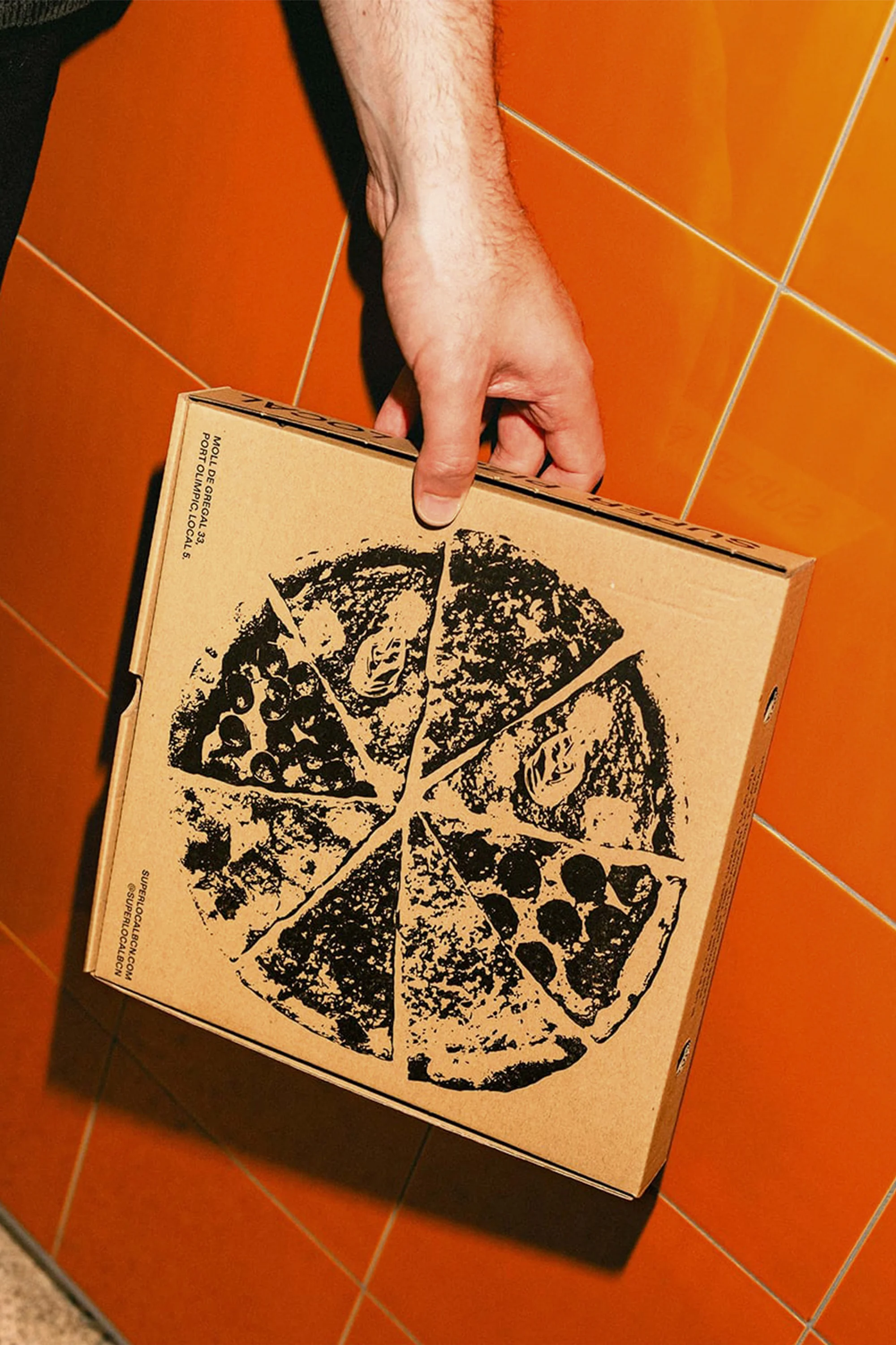 Close-up of a hand holding a square cardboard pizza box with a circular pizza slice design on an orange-tiled background.