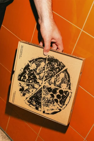 Close-up of a hand holding a square cardboard pizza box with a circular pizza slice design on an orange-tiled background.