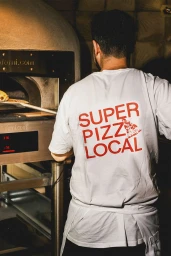 Man wearing a white t-shirt with red Super Pizz Local lettering seen from behind working by a pizza oven