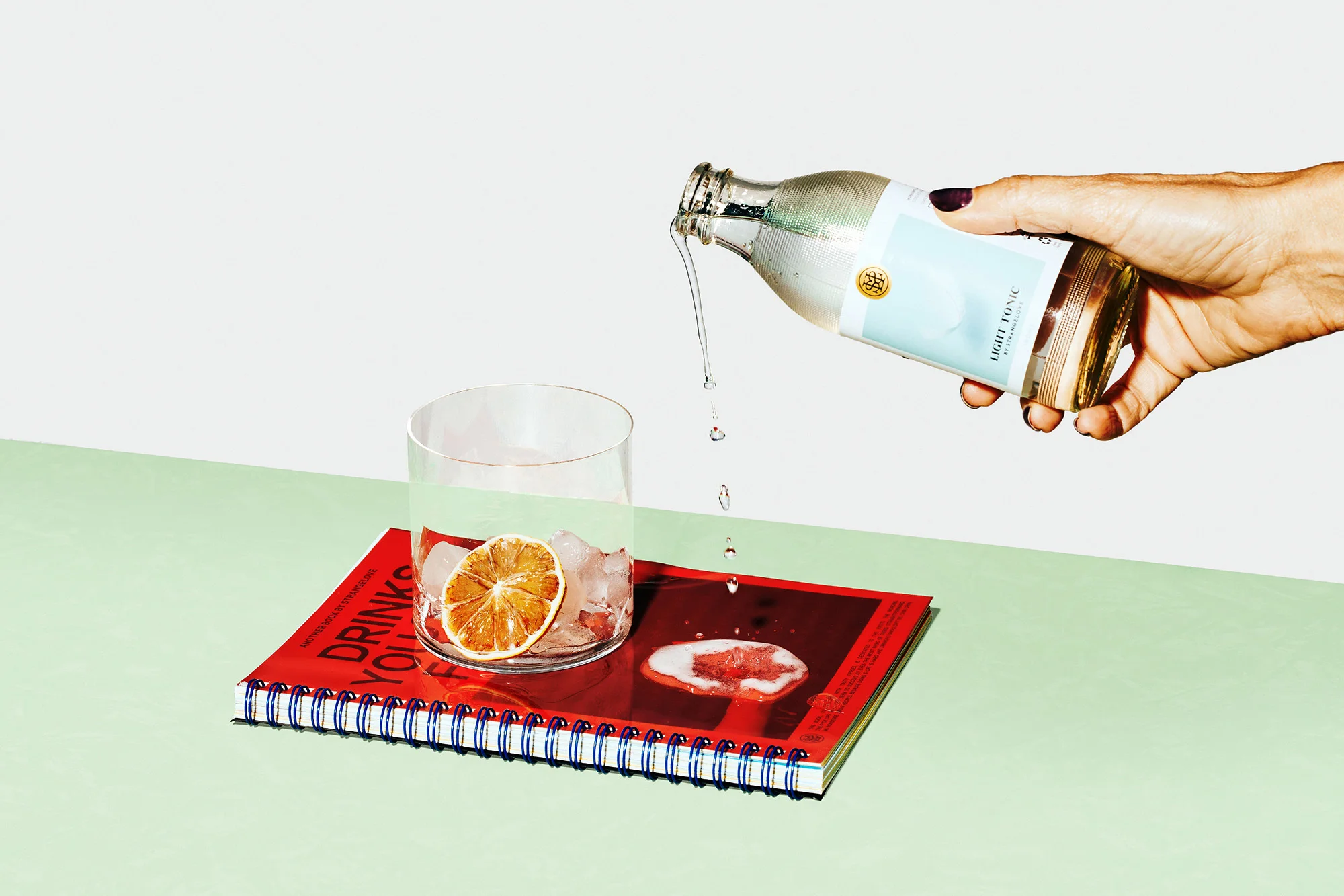 Clear glass filled with ice and a dried orange slice, liquid being poured from a bottle by a person with painted fingernails.