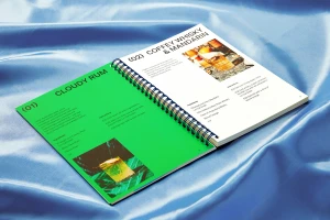Open spiral-bound recipe book resting on a shiny blue satin fabric background with printed cocktail recipes.