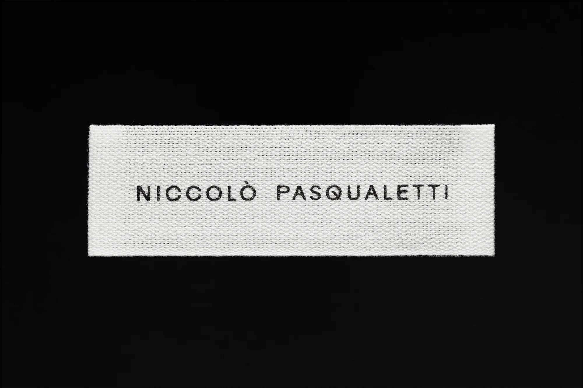Close-up of a white fabric tag with the name NICCOLÒ PASQUALETTI in black, set against a solid black background.