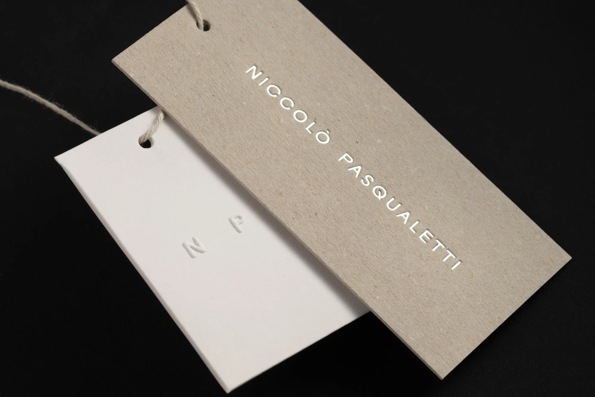 Two tags, one beige with 'Niccolo Pasqualetti' in white letters, and one white with embossed 'NP,' hang against a black background.