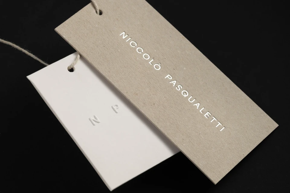 Two tags, one beige with 'Niccolo Pasqualetti' in white letters, and one white with embossed 'NP,' hang against a black background.