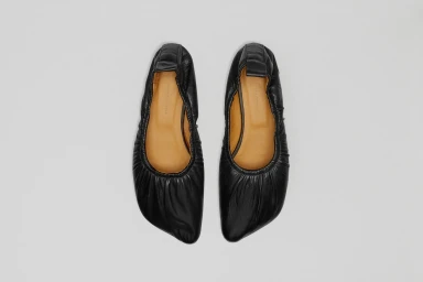 Top-down view of a pair of black leather ballet flats with gathered elastic heels and tan insoles on a neutral grey background