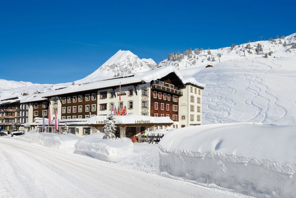 Exterior view of the Hotel Lorunser in Zürs, Austria, showcasing snow-covered landscape, building architecture, and clear blue sky.