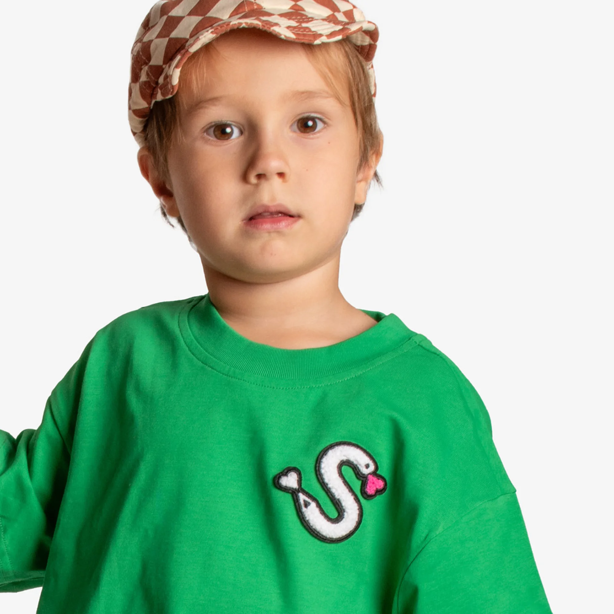 A young boy wearing a green long-sleeved shirt with a snake design and a patterned hat, posing against a plain white background.