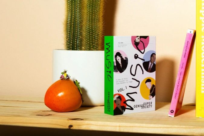 Still life photograph featuring a cactus in a white pot, an orange persimmon with tiny figurines, and music book cover on a wooden shelf.