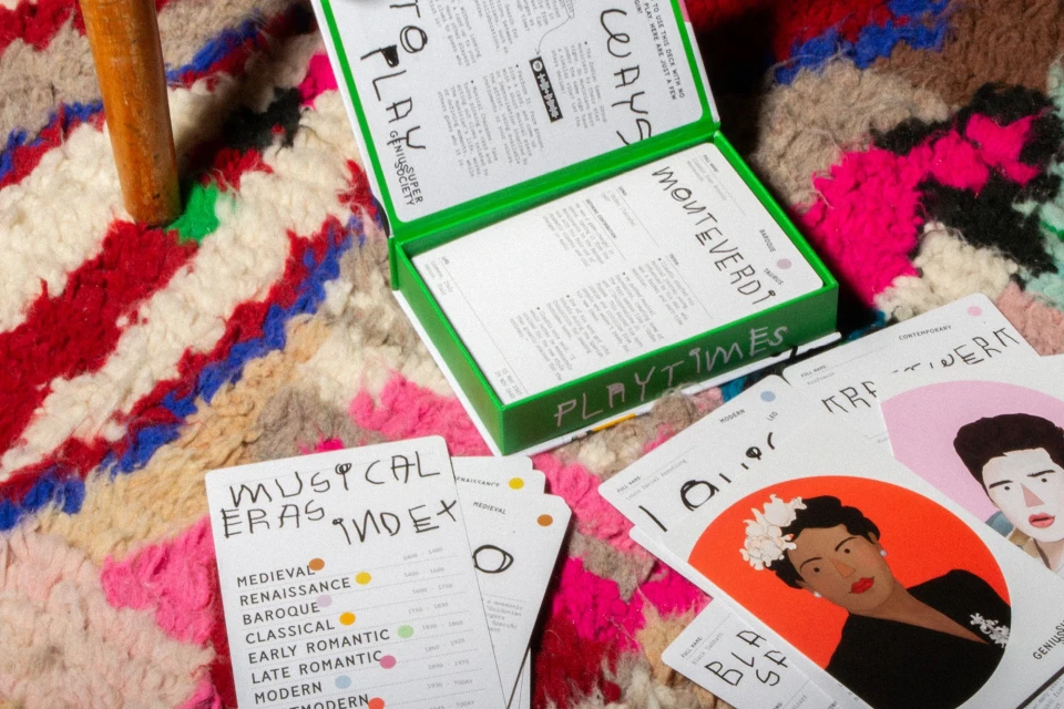 Open green Playtimes Super Genius Society box sits on a colorful striped rug, surrounded by cards with eras and portraits.