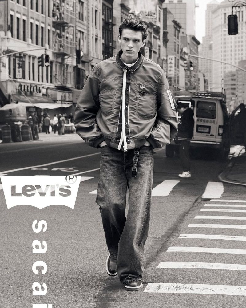 Monochrome outdoor fashion shot of a young adult male model in a bomber jacket and baggy jeans walking on a city street in a street fashion context.
