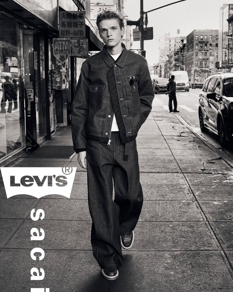 Monochrome portrait of a male model wearing a denim jacket and wide-leg jeans on a city street with buildings and traffic in the background.