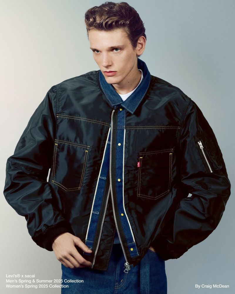 Adult male poses in a fashion editorial shot, wearing a black bomber jacket with denim accents and blue jeans in a studio setting.