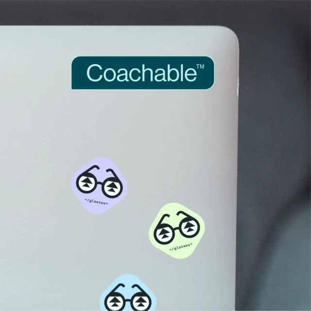 Close-up of a laptop lid with a 'Coachable' sticker and cartoon eyeglasses stickers in various colors and designs.