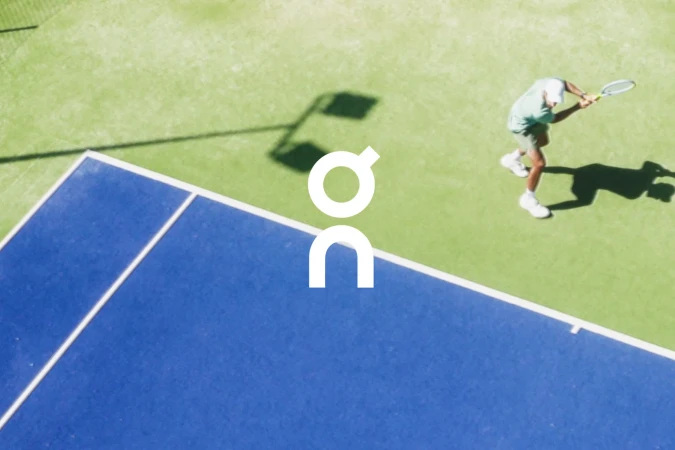 Overhead shot of a tennis player in motion on a green and blue court with the "On" brand logo.