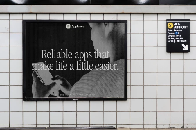 Subway platform displays an ad for 'Applause' featuring a person using a smartphone, alongside a JFK Airport direction sign.